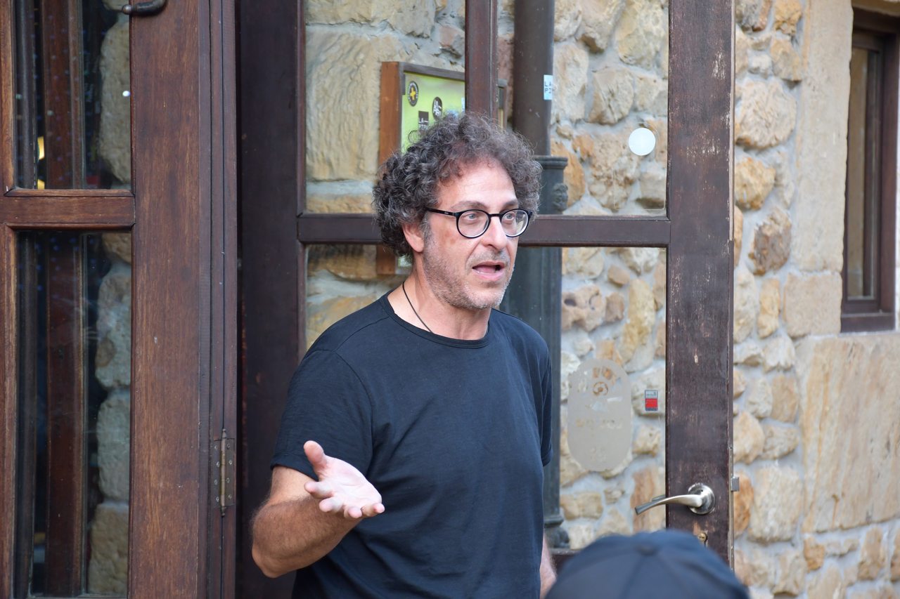 A seated speaker gestures beside a stone doorway at an outdoor gathering
